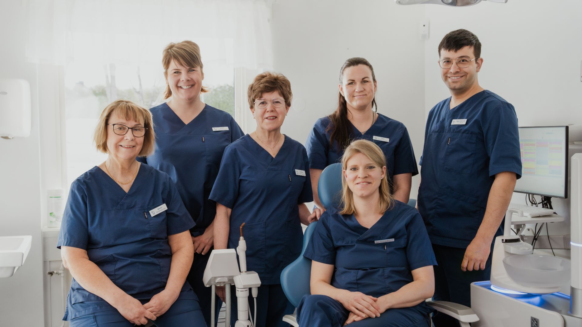 Sex tandläkare i marinblå operationskläder ler tillsammans i en ljus, modern tandläkarmottagning som ägnar sig åt kvalitativ tandvård i Sverige, inklusive implantat.