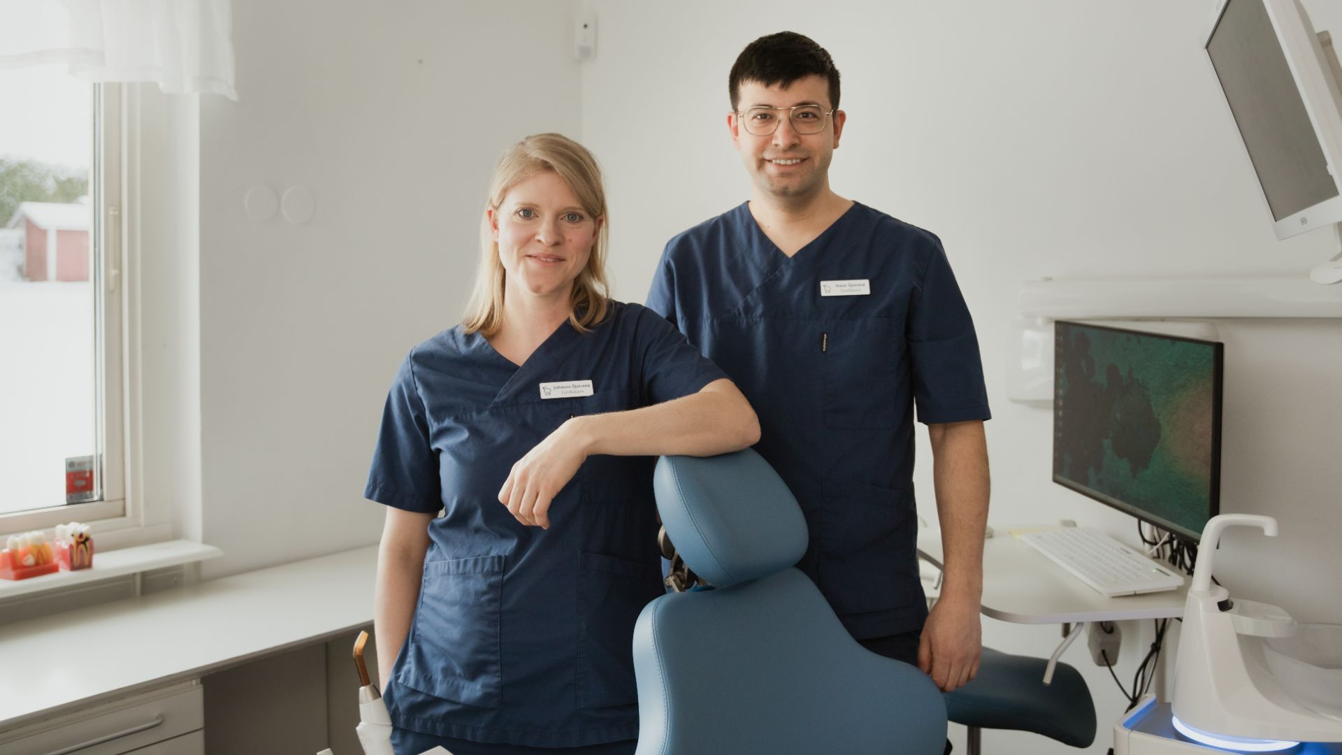 Två tandläkare i marinblå kläder står bredvid en tandläkarstol i en ljus, modern tandvårdsklinik i Årjäng, redo att hjälpa till med dina implantatbehov.
