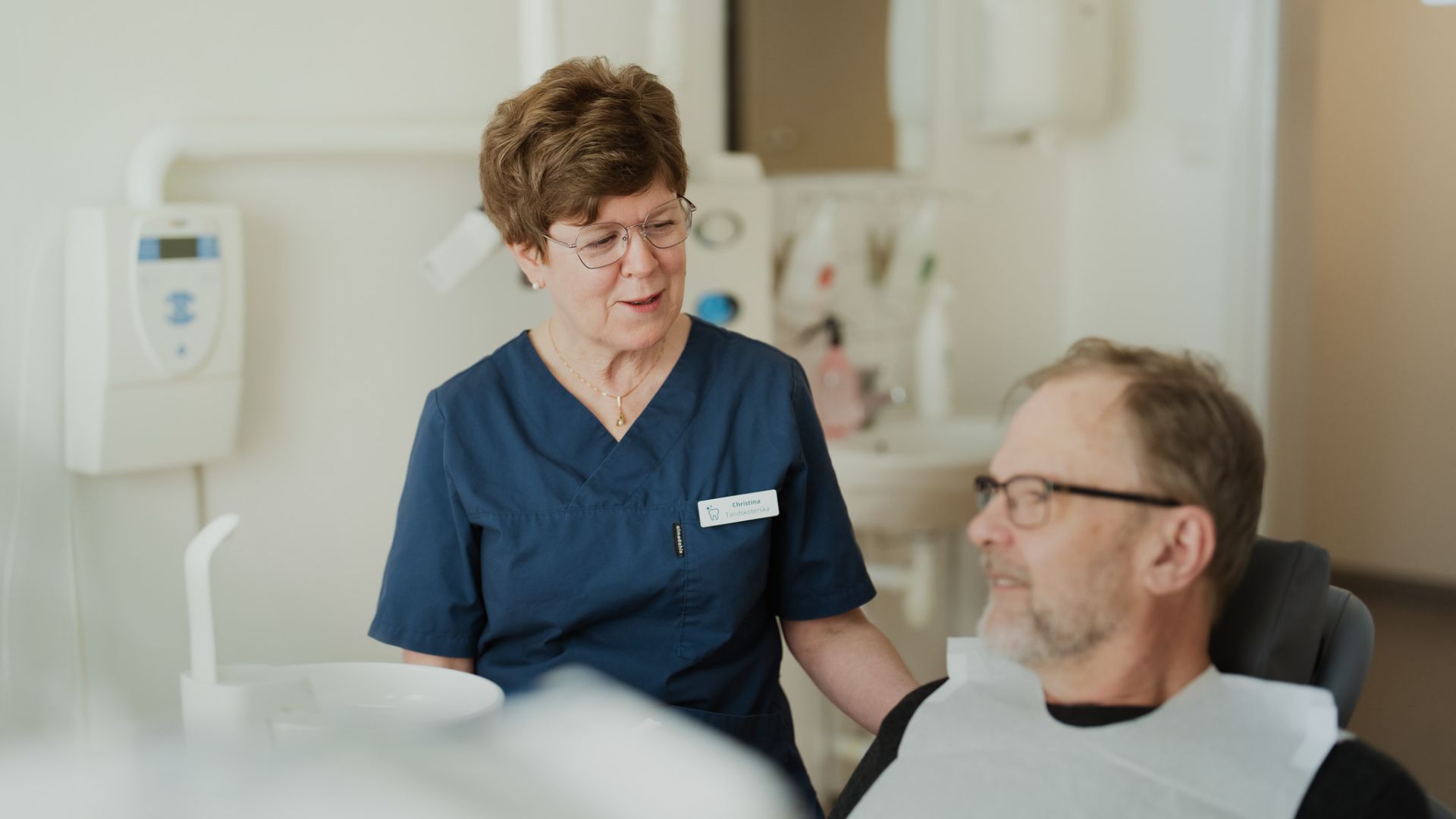 En tandläkare årjäng i blå operationskläder pratar med en sittande manlig patient om implantatmöjligheter på en modern tandläkarmottagning.