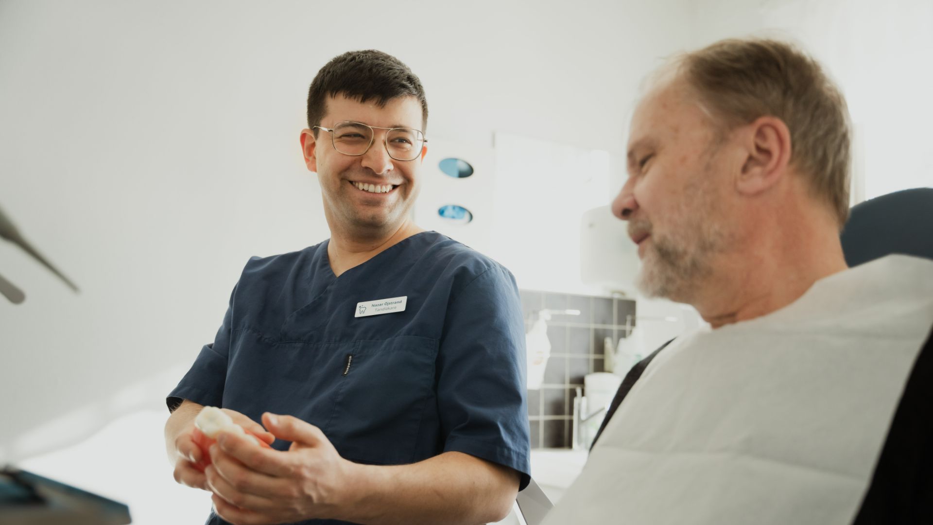 En vårdpersonal i blå operationskläder pratar med en äldre manlig patient med tandläkarhaklapp om tandimplantat i en klinisk miljö.