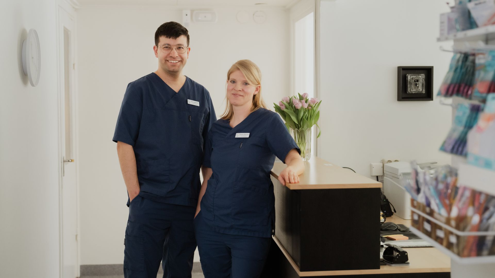Två vårdpersonal i blå operationskläder står vid en receptionsdisk i en ren och väl upplyst tandvårdsklinik i Årjäng, redo att hjälpa patienter som söker expertvård inom tandvård.