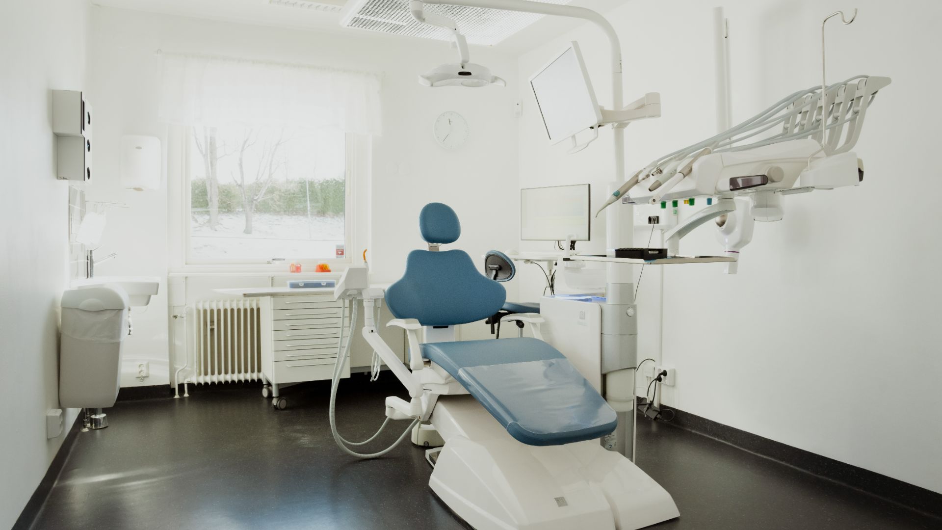 Ett modernt undersökningsrum för tandläkare med en blå tandläkarstol, taklampa, tandläkarinstrument för implantatbehandling, datorskärm och vita väggar.