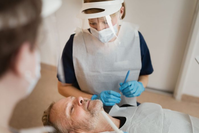 En patient ligger på en tandläkarstol medan två tandläkare Årjäng i masker och ansiktsskydd förbereder sig för ett ingrepp, vilket säkerställer professionell tandvård Årjäng.